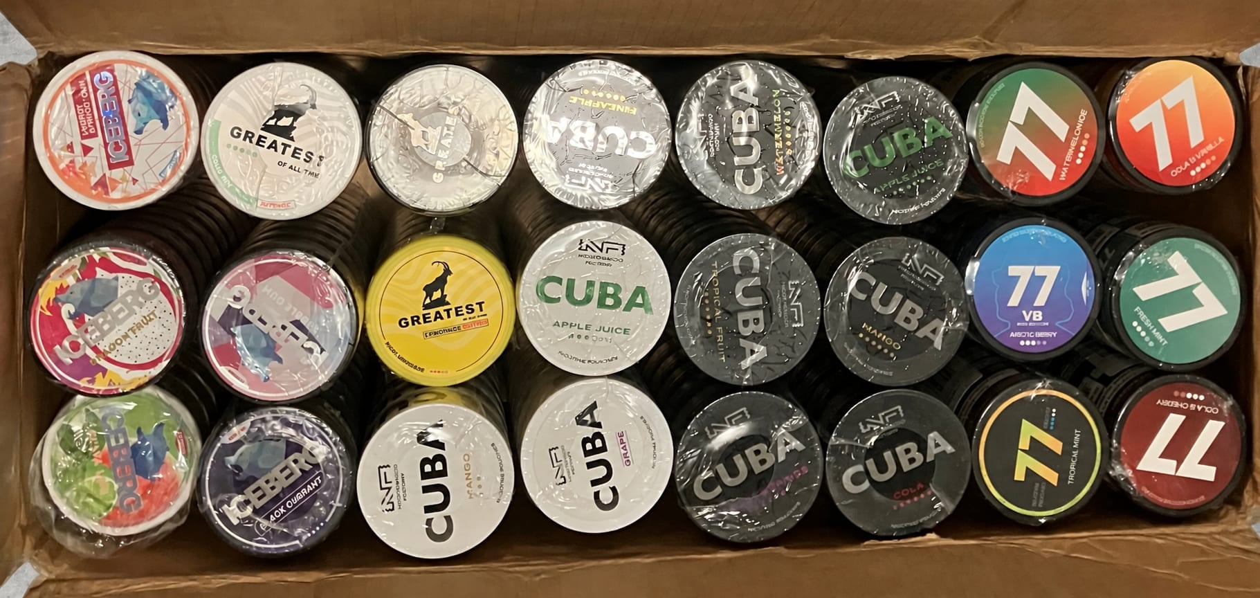 An opened carton of nicotine pouch cans — 77, Cuba, Greatest and Iceberg tins packed in rows.